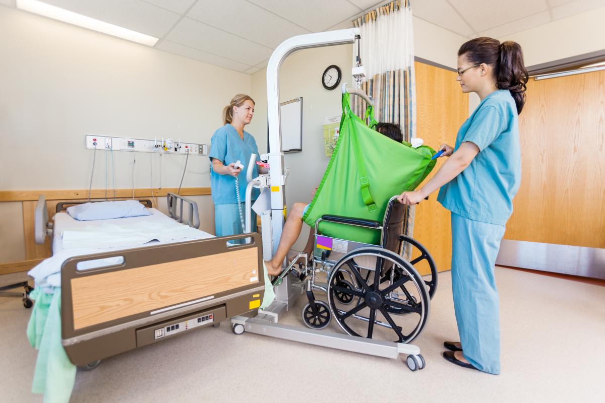 A transfer sling attached to a mobile hoist being operated by two support workers to transfer out of a wheelchair