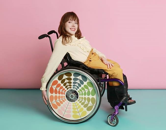 Izzy Keaene poses against a light blue and pink backdrop in her decorated wheelchair, which has a colorful leaf design on the wheel