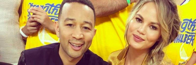 John Legend and Chrissy Teigan sit together smiling at a sports event