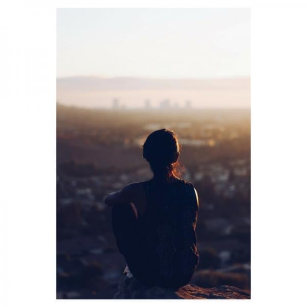 Woman sits at the edge of a cliff alone, staring out at a city
