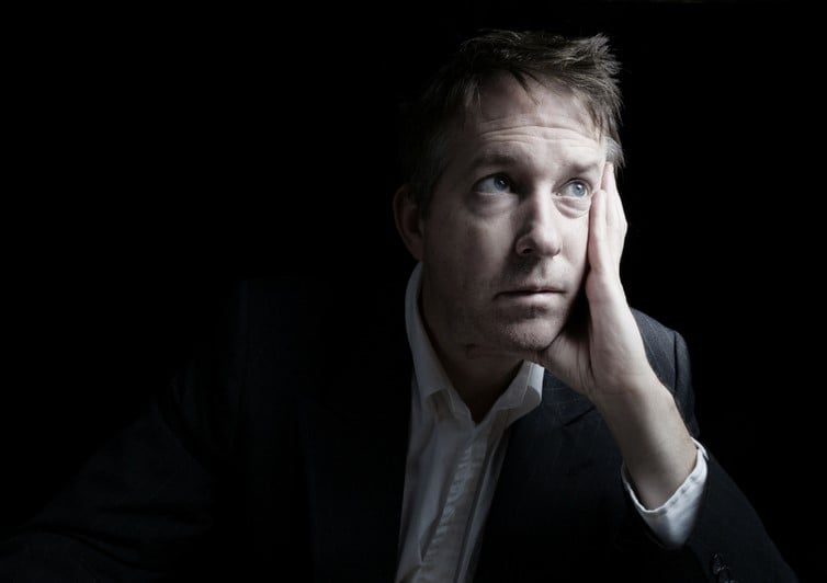 Man in a suit sits with a sad expression on his face, which is resting in his hand