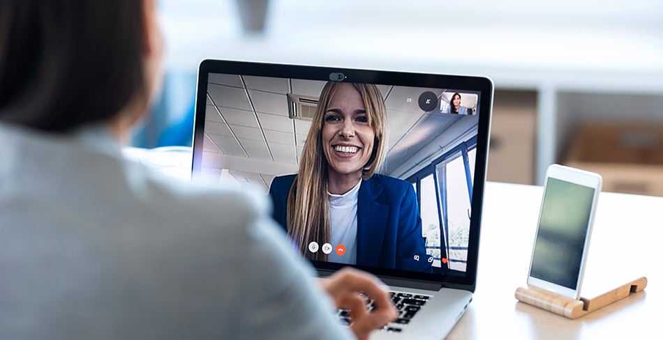 Two people having a metting online. We are looking over the shoulder of one of them and see a smiling woman on the laptop screen in front of them