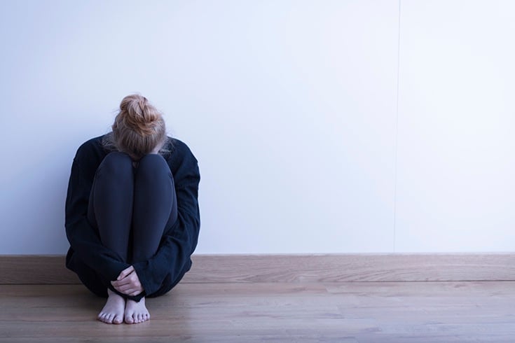 a woman sits on the floor against a white wall in the corner of a photo with her head on her knees and arms clutching her legs