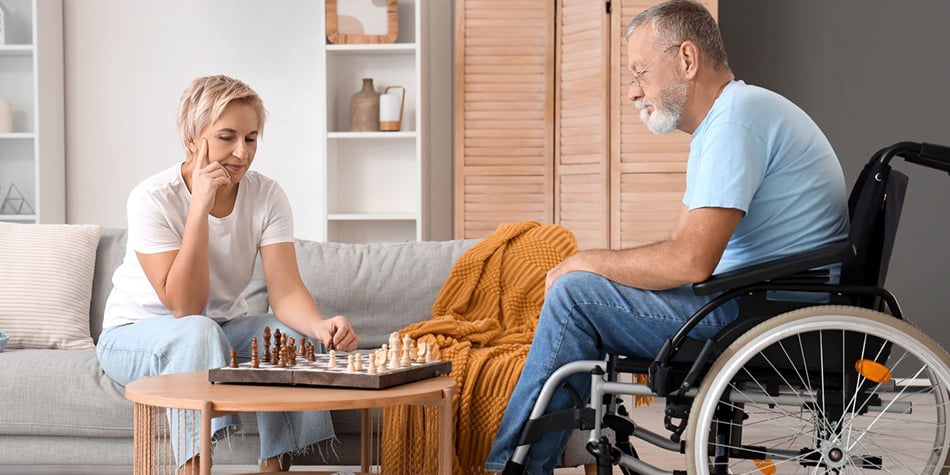 Man seated in wheelchair playing chess with woman seated on couch